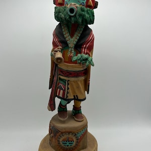 May include: A carved wooden Kachina doll. The figure has a turquoise face, a black headdress with feather accents, and is wearing a red and black robe. It holds a bell and a sprig of leaves. The doll stands on a wooden base.