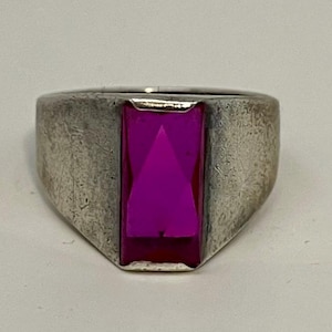 May include: A silver-toned ring with a large, rectangular, magenta-colored gemstone. The ring has a wide band and a slightly worn appearance, suggesting it is vintage or antique. The gemstone is faceted, catching the light.