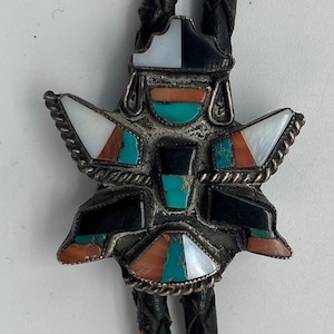 May include: A silver bolo tie with a geometric design featuring turquoise, coral, and mother-of-pearl stones. The bolo tie is on a black leather cord.