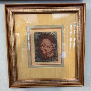 May include: Framed artwork featuring a portrait of an elderly person with glasses. The artwork is in a gold-colored frame with a light-colored mat. The portrait is in shades of red, brown, and blue, with the text "'Gurneshaut'" at the bottom.