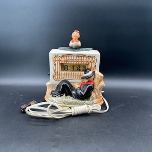 May include: A ceramic clock with a digital display showing 8:12. The clock is shaped like a small house with a man sitting on the porch. The man is wearing a hat and a jacket. There is a small figure of a person on top of the house.