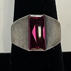 May include: A silver-toned ring with a rectangular, deep red gemstone. The ring has a wide band and a textured surface, with the gemstone set prominently in the center. The gemstone reflects light, creating a vibrant appearance.