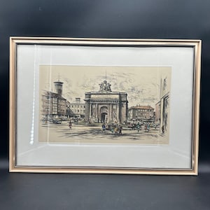 May include: A framed black and white drawing of a city scene with a large archway in the center. The archway has a statue on top and is surrounded by buildings. There are people walking in the street.