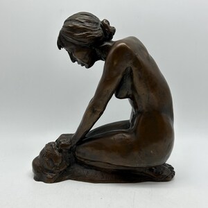May include: A bronze sculpture of a nude woman kneeling with her head bowed. The sculpture is detailed and realistic, with a smooth, polished finish.
