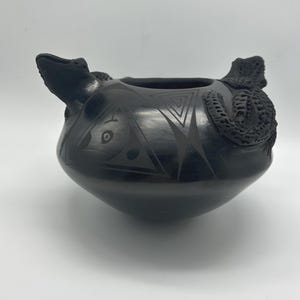 May include: Black ceramic pottery bowl with a rounded base and two sculpted reptile handles. The bowl features etched geometric designs and a smooth, glossy finish. A unique decorative piece.