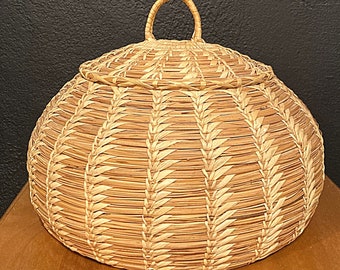 May include: A round, woven basket with a lid and a handle. The basket is made of natural materials and has a light brown color.