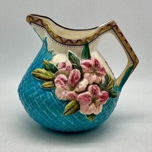 May include: A blue and white ceramic pitcher with a floral design. The pitcher has a handle and a wide mouth. The flowers are pink and white and have green leaves. The pitcher is decorated with a gold trim.