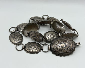 May include: A silver-toned belt with oval and rounded rectangular links. Each link features a geometric design. The belt has a large oval buckle with a detailed pattern. The belt is likely made of metal and is displayed on a white background.