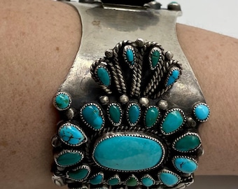 May include: A silver cuff bracelet with turquoise stones. The bracelet features a large oval turquoise stone in the center, surrounded by smaller turquoise stones. The bracelet has a decorative design with a floral element at the top.