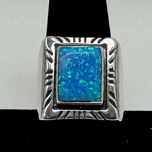 May include: A silver ring with a rectangular, blue-green opal stone. The stone is set within a decorative frame. The ring has a wide band and a detailed design around the stone, showcasing a classic jewelry style.