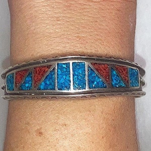 May include: A silver bracelet with turquoise and red coral inlay. The bracelet has a geometric design with alternating turquoise and coral sections.