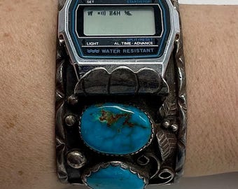 May include: A silver-toned Timex digital watch with a black and blue face, featuring the text "LITHIUM QUARTZ" and "WATER RESISTANT". The watch is set in a wide, ornate silver cuff bracelet with two oval turquoise stones and decorative floral elements.