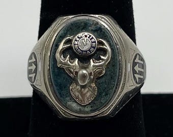 Könnte beinhalten: Silberring mit rechteckiger Fassung mit einem grünen Stein. Der Ring hat einen Hirschkopf und ein kreisförmiges Emblem mit Text. Die Seiten des Rings weisen Pfeildesigns auf.