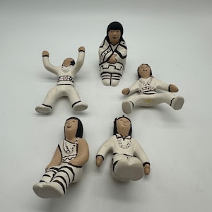 May include: Five hand-painted ceramic figurines of Native American people. The figurines are all wearing traditional clothing and have black hair. They are all sitting or standing in different poses.