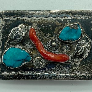 May include: A silver belt buckle with turquoise and coral stones. The buckle is decorated with a floral design and has a textured surface.