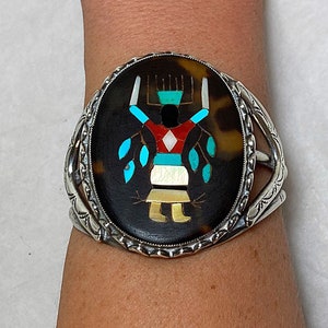 May include: A silver bracelet with a large oval-shaped inlay featuring a stylized Native American figure in a colorful mosaic of turquoise, red, white, and black stones. The figure is surrounded by a brown tortoiseshell-like background.
