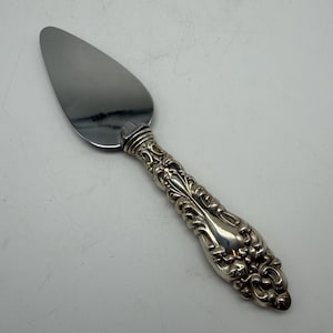May include: A silver-toned cake server featuring a smooth, triangular metal blade and an ornate, embossed handle with intricate scrollwork and floral-inspired detailing. The handle has a polished finish, while the blade is reflective and plain.