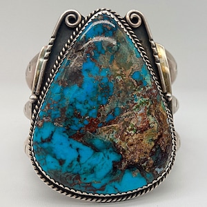 May include: A silver and turquoise Native American bracelet with a large, irregularly shaped turquoise stone set in the center. The stone has a blue, green, and brown color pattern.