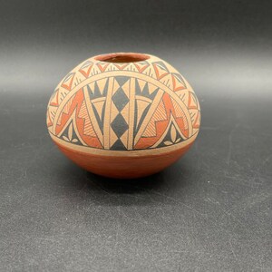 May include: A small, round, terracotta pottery vase with a geometric design in black, white, and orange. The vase has a wide opening at the top.