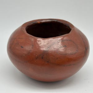 May include: A round, reddish-brown ceramic bowl with a wide opening. The bowl has a smooth, glossy finish with darker brown markings and a dark interior. The pottery appears handmade.