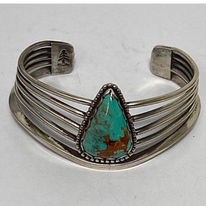 May include: A silver bracelet with a turquoise stone set in the center. The bracelet has five bands and a tree design on the back.