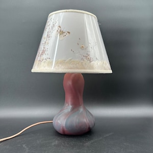 May include: A decorative table lamp with a pink and gray base and a white lampshade. The lampshade features a butterfly and floral design. The lamp has a cord for plugging into an electrical outlet.
