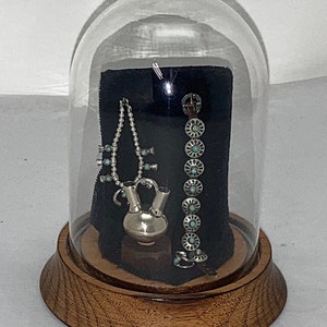 May include: A glass dome display case with a wooden base, showcasing a silver necklace with turquoise stones, a silver bracelet with turquoise stones, and a silver pitcher with a turquoise stone accent.