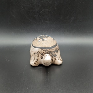 May include: A ceramic turtle figurine with black and white painted designs. The turtle is in a walking position with its head and legs visible.