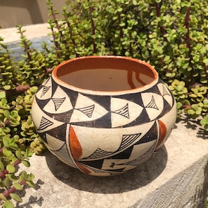 May include: A round ceramic bowl featuring a cream-colored base with a geometric pattern of black triangles and orange accents. The interior rim is painted in a solid orange color, and the exterior surface displays a repeating hand-painted design.