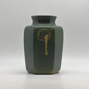 May include: A matte dark green ceramic vase with a hexagonal body and a short, wide neck. The front surface features a singular, raised, light brown organic drip-like glaze accent that extends downward from a circular, textured embossed detail.