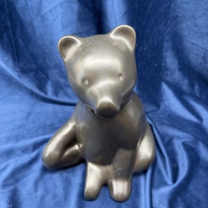 May include: A metallic dark gray ceramic bear figurine in a seated position. The bear has a smooth, reflective surface, rounded ears, and a simple facial structure with subtle indentations for the eyes and a prominent, rounded snout.