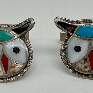 May include: A pair of silver cufflinks featuring owl designs. Each owl has a white face with black eyes, a red beak, and a turquoise or green head. The wings are black, and the body has a shell-like pattern.