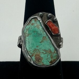 May include: A silver ring with a turquoise and coral stone inlay. The ring has a unique, organic shape and is set with a large, green turquoise stone. The coral stone is a bright red and is set in the center of the ring.