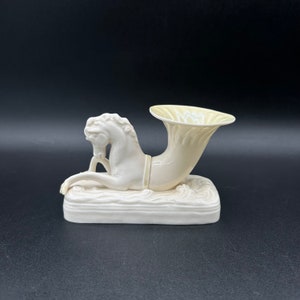 May include: A white porcelain figurine of a horse with a yellow horn-shaped vase. The figurine is on a rectangular base with a wave-like design.