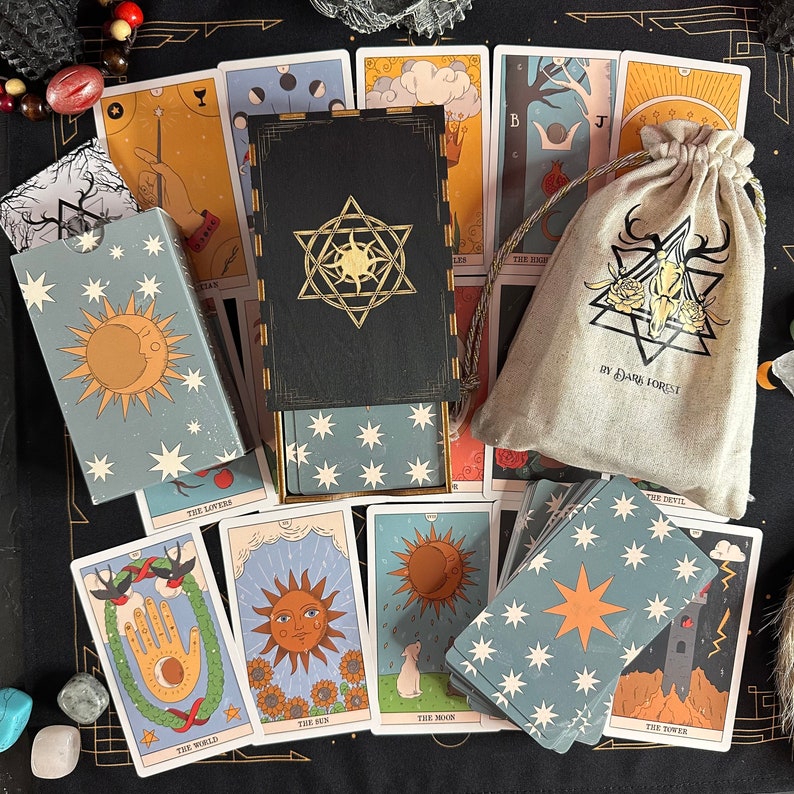 K&ouml;nnte beinhalten: Ein Tarot-Kartenset bestehend aus einer schwarzen Holzschachtel mit goldenem Sonnenemblem, einem beigen Kordelzugbeutel mit Totenkopf- und Blumenmotiv sowie der Aufschrift "BY DARK FOREST" und mehreren Karten mit Sonnen-, Mond- und Naturmotiven.