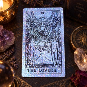 May include: A rectangular tarot card featuring a holographic, glittery finish with black line art depicting The Lovers. The card shows two figures standing beneath a large winged angel figure, with the Roman numeral VI at the top and the text THE LOVERS.