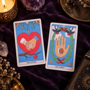 K&ouml;nnte beinhalten: Zwei Tarotkarten auf einer violetten Samtoberfl&auml;che. Die linke Karte mit der Aufschrift THE LOVERS zeigt zwei ineinander verschlungene H&auml;nde in einem gro&szlig;en roten Herzen. Die rechte Karte, THE WORLD, zeigt eine Hand mit Symbolen und Kranz.