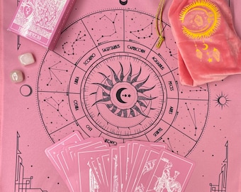Puede incluir: Tela de tarot rosa con un diseño de rueda zodiacal y una bolsa de terciopelo rosa. La tela presenta un diseño de sol y luna negros en el centro. La bolsa tiene un diseño de sol y luna dorados. La tela está rodeada por un borde negro con un diseño geométrico. La tela está colocada sobre una mesa con una baraja de cartas de tarot.