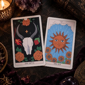 May include: Two tarot cards featuring The Devil and The Sun. The Devil card displays a horned skull, red roses, and flames on a black background. The Sun card shows a radiant sun face above sunflowers on a light blue background with white clouds.