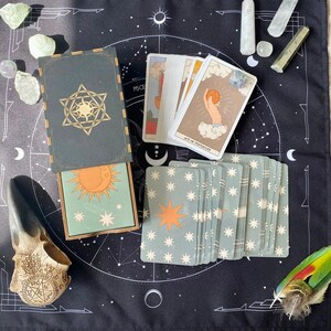 K&ouml;nnte beinhalten: Ein Tarot-Kartendeck mit einer dunkelgr&uuml;nen Schachtel, die ein goldenes Stern- und Sonnenemblem zeigt, einer teilweise ge&ouml;ffneten Schachtel mit einer Sonnenillustration und mehreren ausgebreiteten Karten. Die Karten zeigen Himmelsmotive.