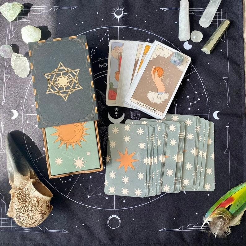 K&ouml;nnte beinhalten: Ein Tarot-Kartendeck mit einer dunkelgr&uuml;nen Schachtel, die ein goldenes Stern- und Sonnenemblem zeigt, einer teilweise ge&ouml;ffneten Schachtel mit einer Sonnenillustration und mehreren ausgebreiteten Karten. Die Karten zeigen Himmelsmotive.