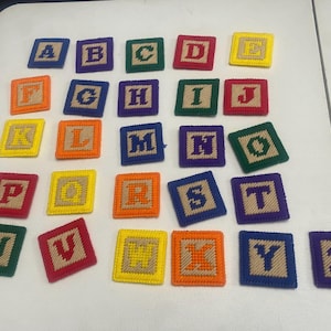 May include: A collection of colorful, square alphabet blocks. Each block features a letter from A to Z, stitched in various colors like red, yellow, green, and blue. The blocks are arranged on a white surface, ideal for educational play.
