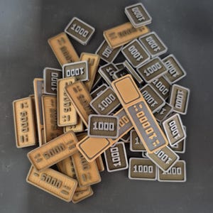 Op de afbeelding: Een verzameling rechthoekige spel fiches, voornamelijk beige en grijs, met de nummers "1000" en "5000" erop gedrukt. Sommige fiches tonen ook "10000". De fiches hebben een getextureerd oppervlak.