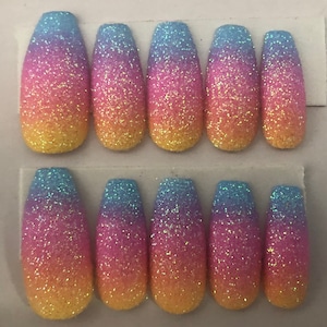 May include: Ten long, oval-shaped, ombre glitter press-on nails. The nails are a gradient of blue, pink, and yellow.