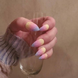 May include: Close-up of a hand with long, pointed nails painted with a gradient of pink, yellow, and blue glitter.