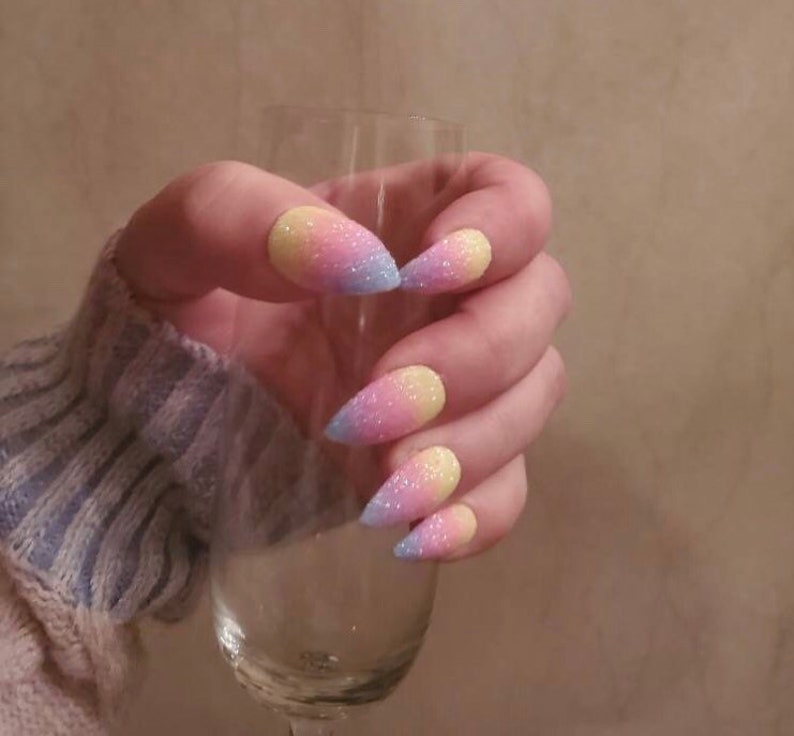 May include: Close-up of a hand with long, pointed nails painted with a gradient of pink, yellow, and blue glitter.