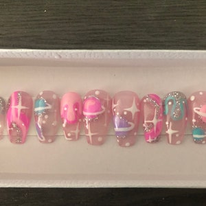 Puede incluir: Conjunto de diez uñas postizas rosas y blancas con un diseño de temática espacial. Las uñas presentan estrellas blancas, planetas y remolinos.