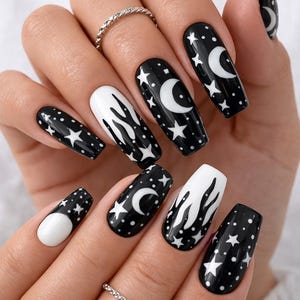 Puede incluir: Una mano muestra uñas artificiales largas en forma de ataúd. Las uñas están pintadas en blanco y negro, con diseños celestiales como lunas, estrellas y patrones de llamas. Un anillo plateado adorna un dedo.