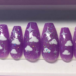 Puede incluir: Un juego de diez uñas postizas moradas con acabado brillante. Cada uña presenta diseños de nubes y estrellas blancas, con acentos de purpurina iridiscente. Las uñas están dispuestas en fila.