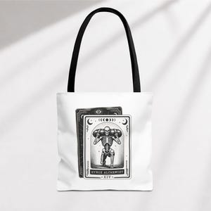 May include: White tote bag with black handles featuring a black and white tarot card design. The card depicts a skeleton figure carrying a weight, with the text "HYROX ALCHEMIST - XIV." Another card is visible behind it.